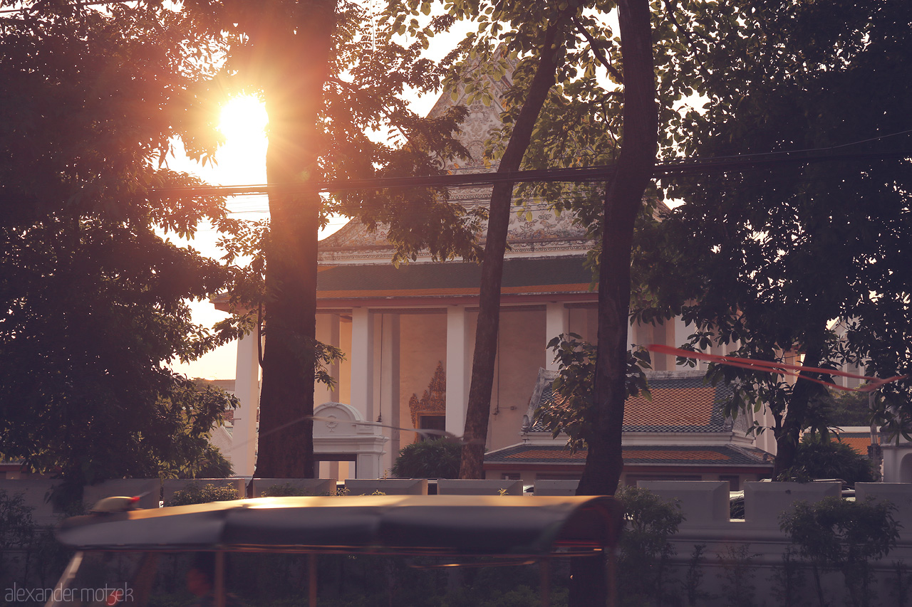 Sunlit Serenity in Ban Bat Foto von A tranquil moment in Ban Bat, Bangkok as the sun sets behind a temple. The peaceful atmosphere is accentuated by the silhouette of lush trees.