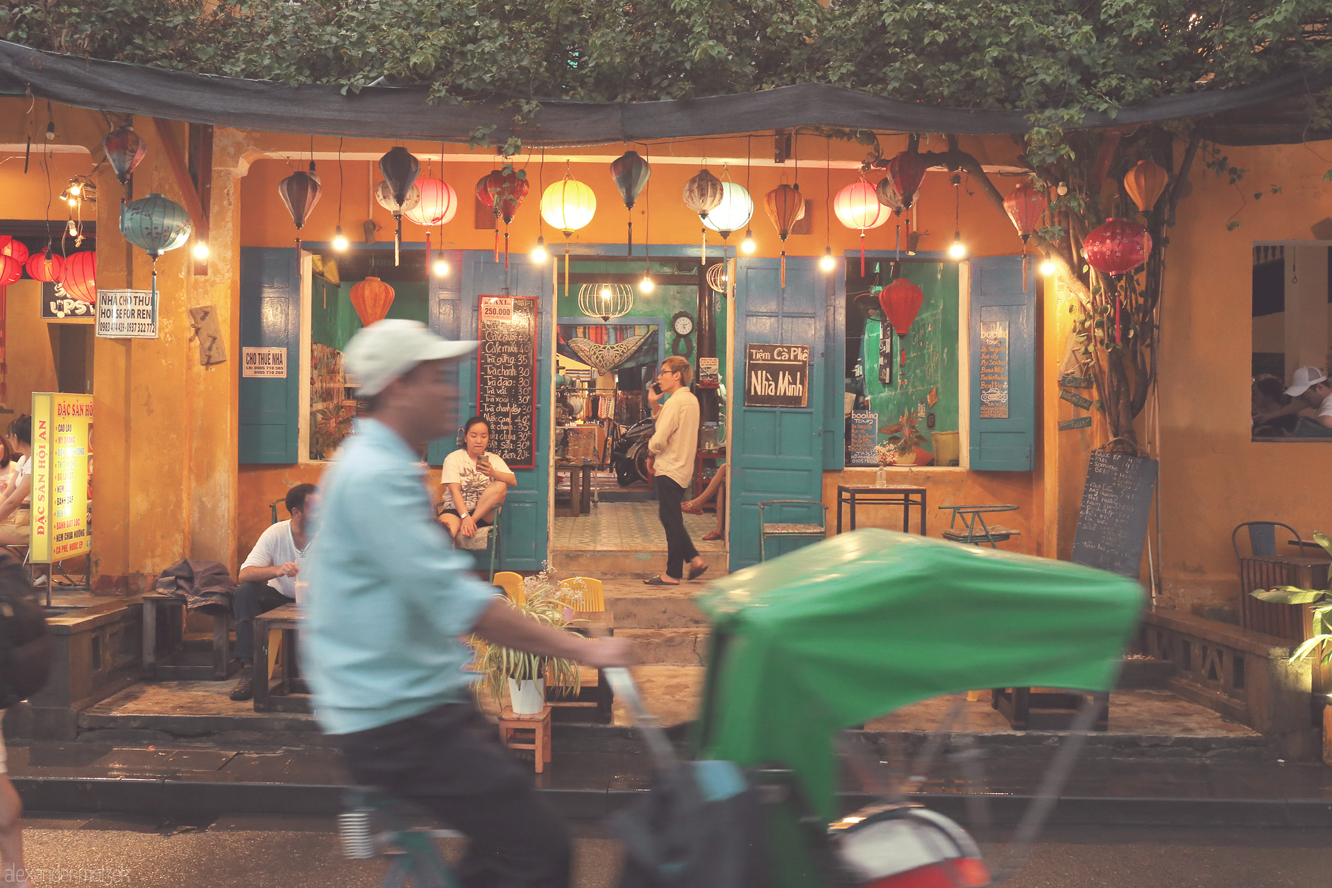 Foto von A vibrant Hoi An café glows beneath colorful lanterns as a cyclo glides past, capturing the city's charm and evening bustle in Vietnam.