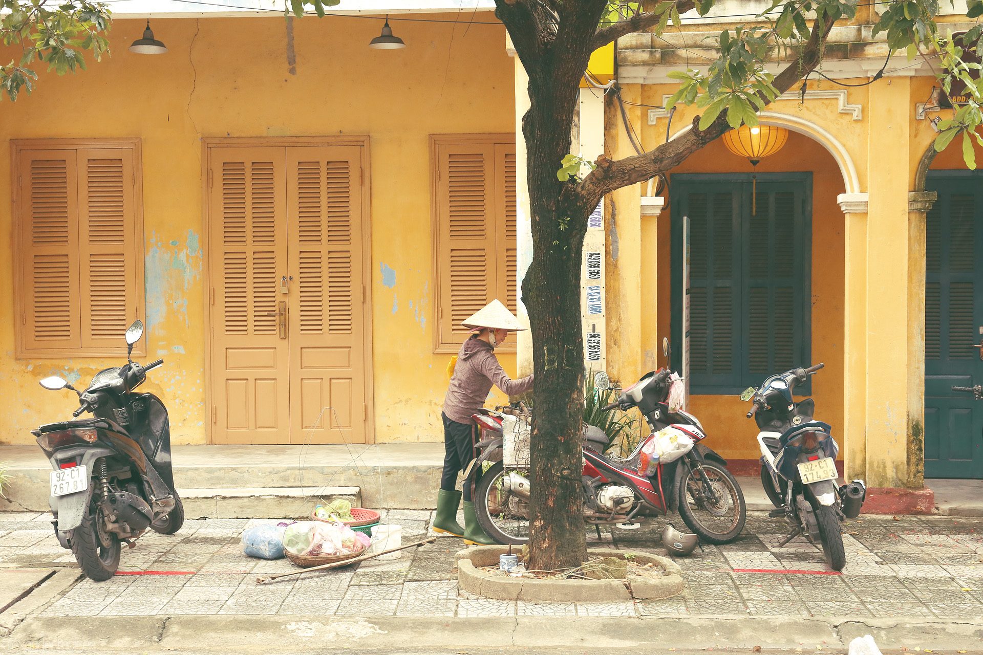 Foto von A woman in a nón lá hat, street vendors’ baskets, and scooters beneath Hoi An’s golden walls and lanterns. Morning unfolds, Vietnam style.