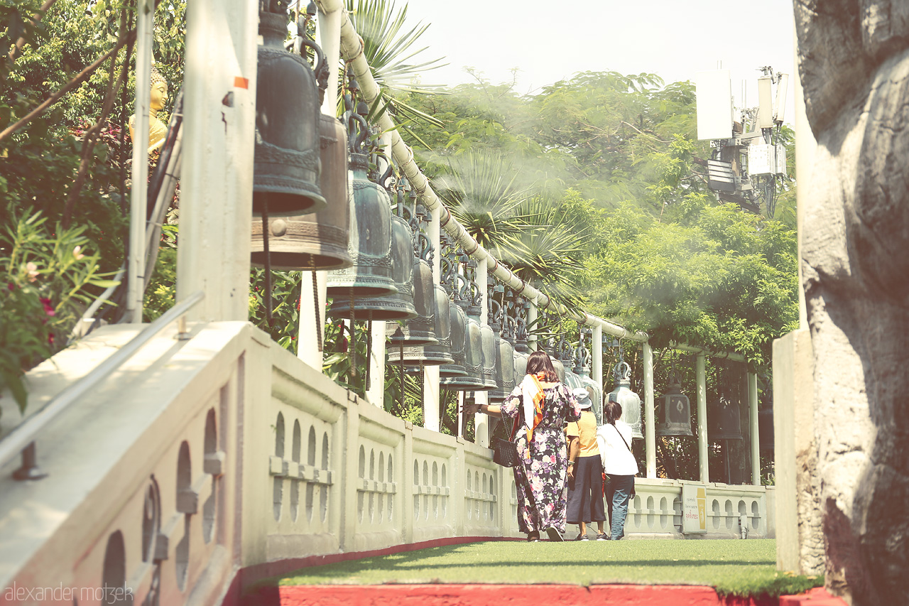 Foto von In Ban Bat, Bangkok, visitors stroll by a tranquil row of bells, capturing a serene mix of tradition and nature.