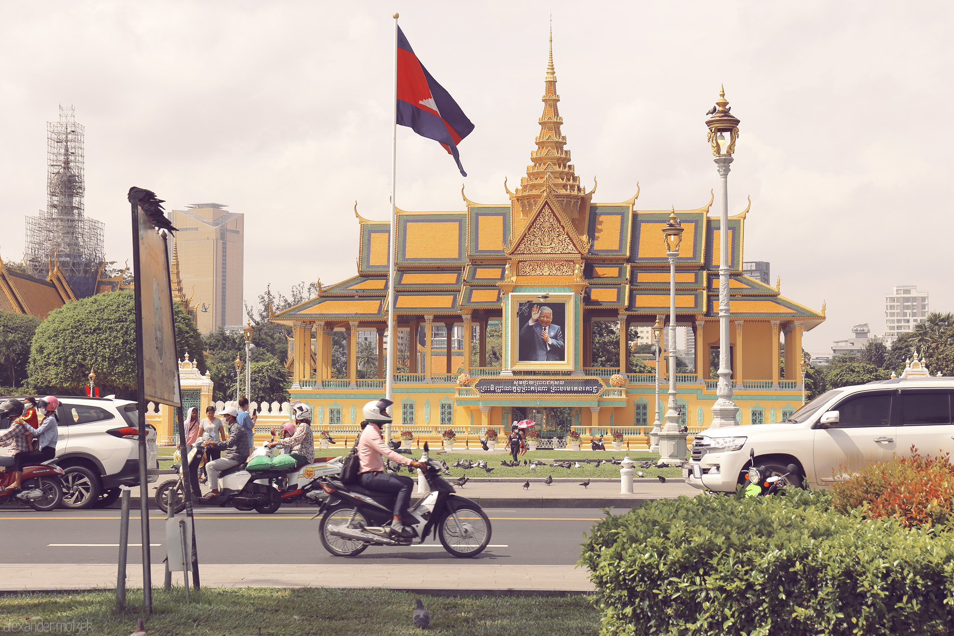 Foto von Beneath the royal gold of Phnom Penh, daily life streams by—scooters, locals, and pigeons beneath the Cambodian flag fluttering in the sun.