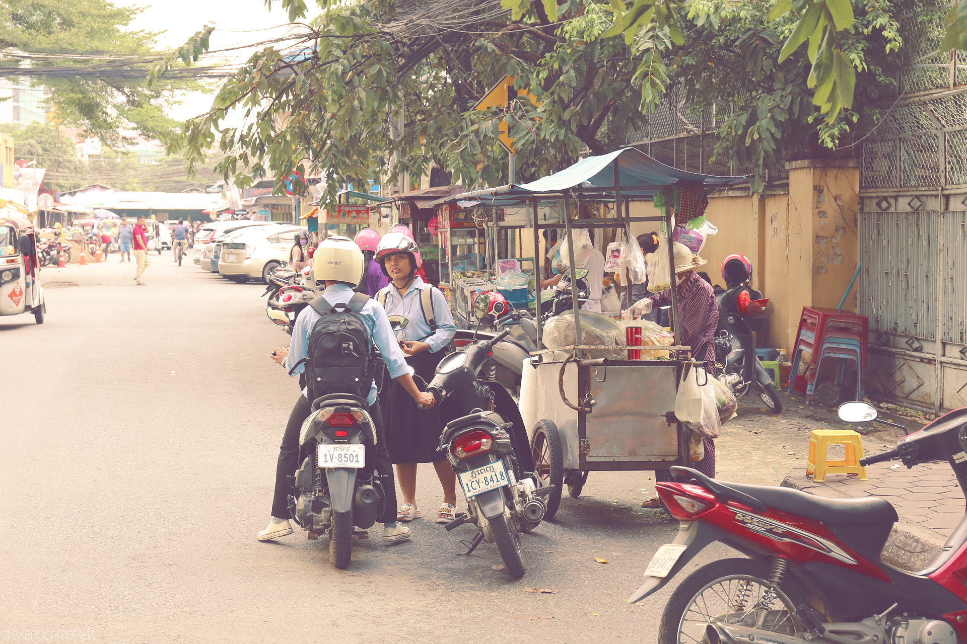 Sabay Street Scene Foto von Local life unfolds on a Phnom Penh street: scooters, school uniforms, and a bustling food cart beneath lush trees in Cambodia's capital.