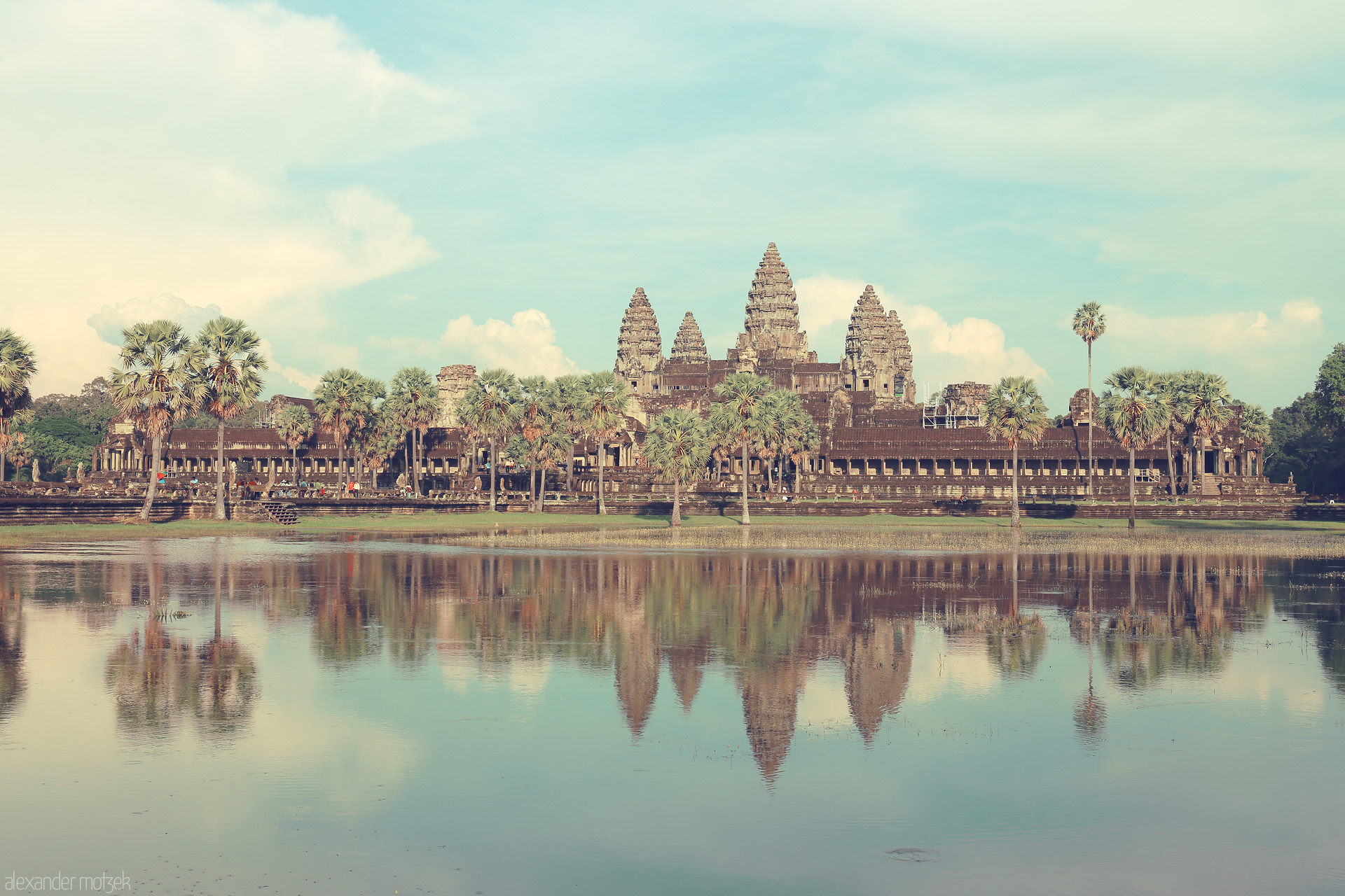 Khmer Elysium Reflections Foto von The iconic spires of Angkor Wat cast a dreamy reflection over tranquil waters in Siem Reap, Cambodia, framed by swaying palm trees.