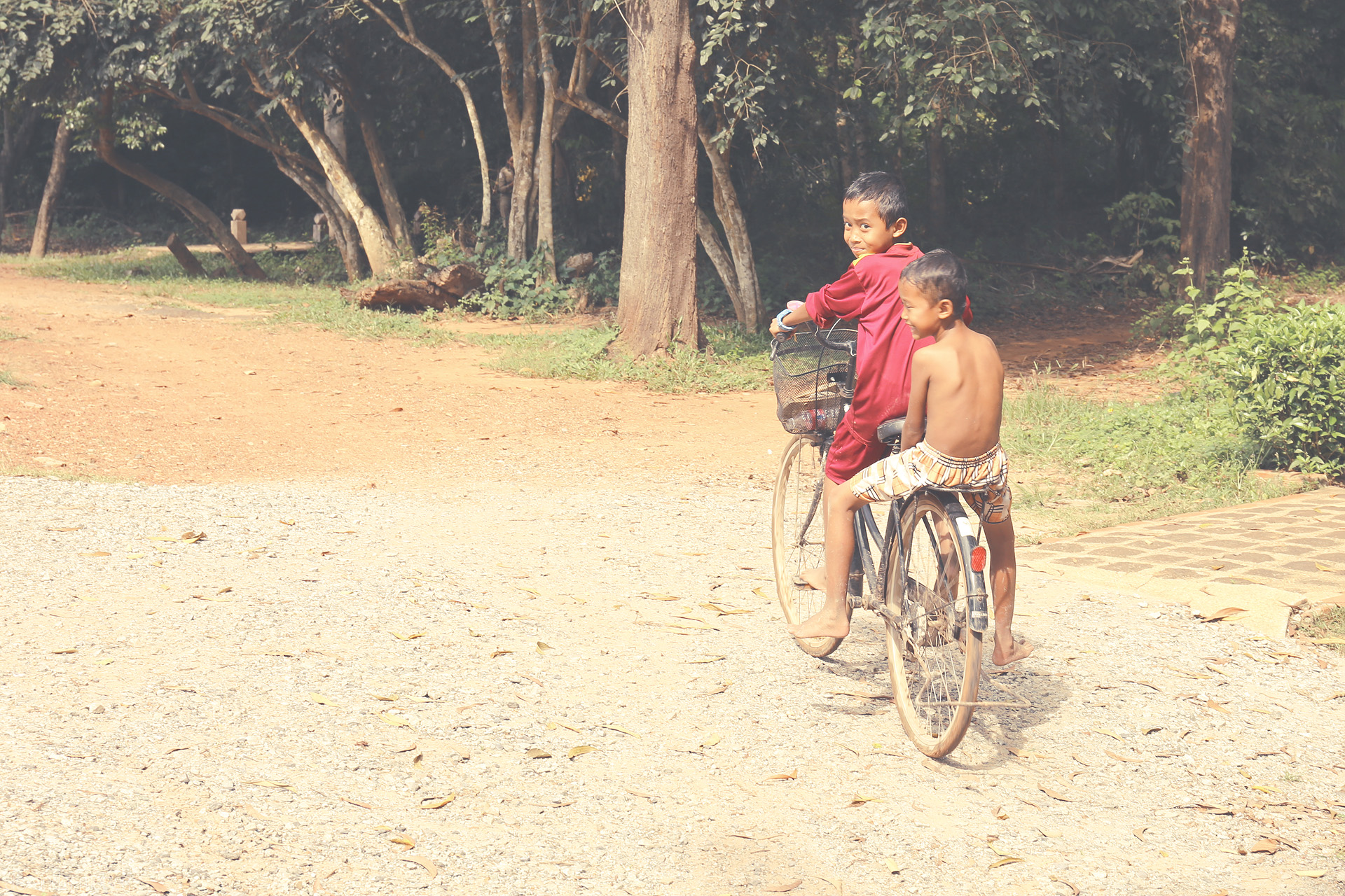 Smiles on Srok Khmer Roads Foto von Two young boys share a bike, laughter, and a sunlit path near Angkor Wat, Siem Reap—where Khmer innocence and adventure meet.