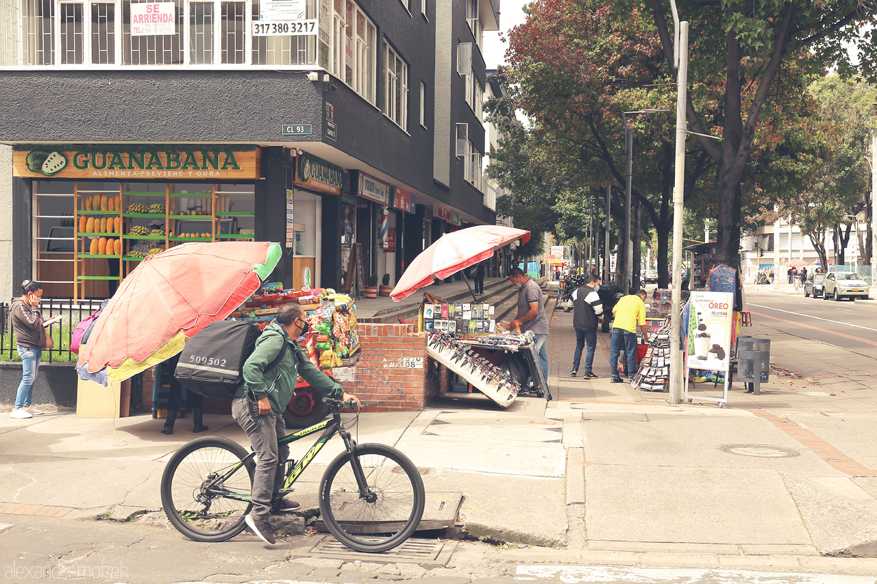 Foto von A bustling street scene in Chapinero, Bogotá. A cyclist passes by local vendors under vibrant umbrellas, capturing the dynamic spirit of urban Colombia.