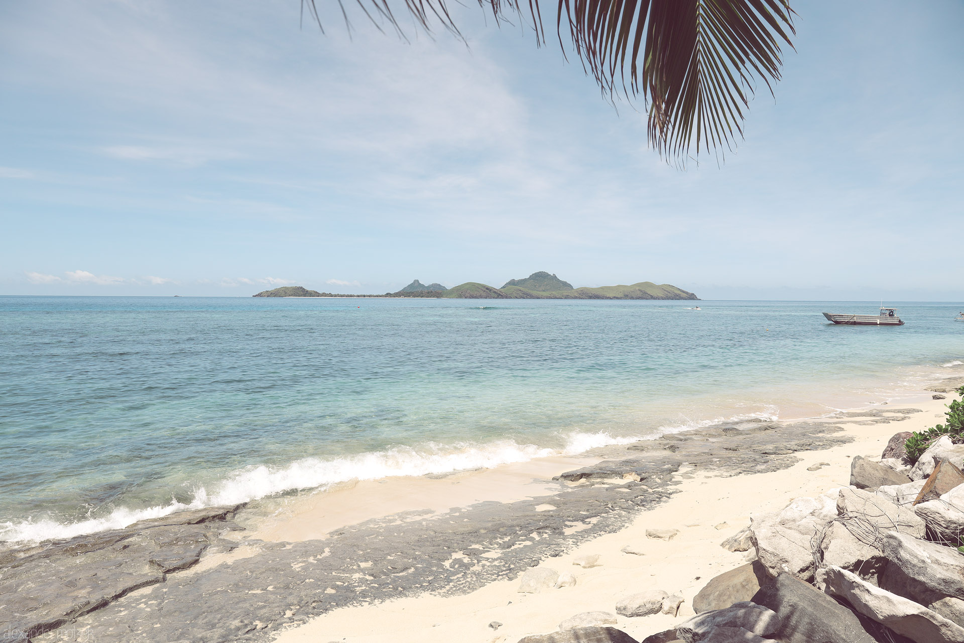 Foto von Palm-framed beach with soft surf, rocky sand, a small boat, and distant green islets on the horizon in Tokoriki, Fiji.