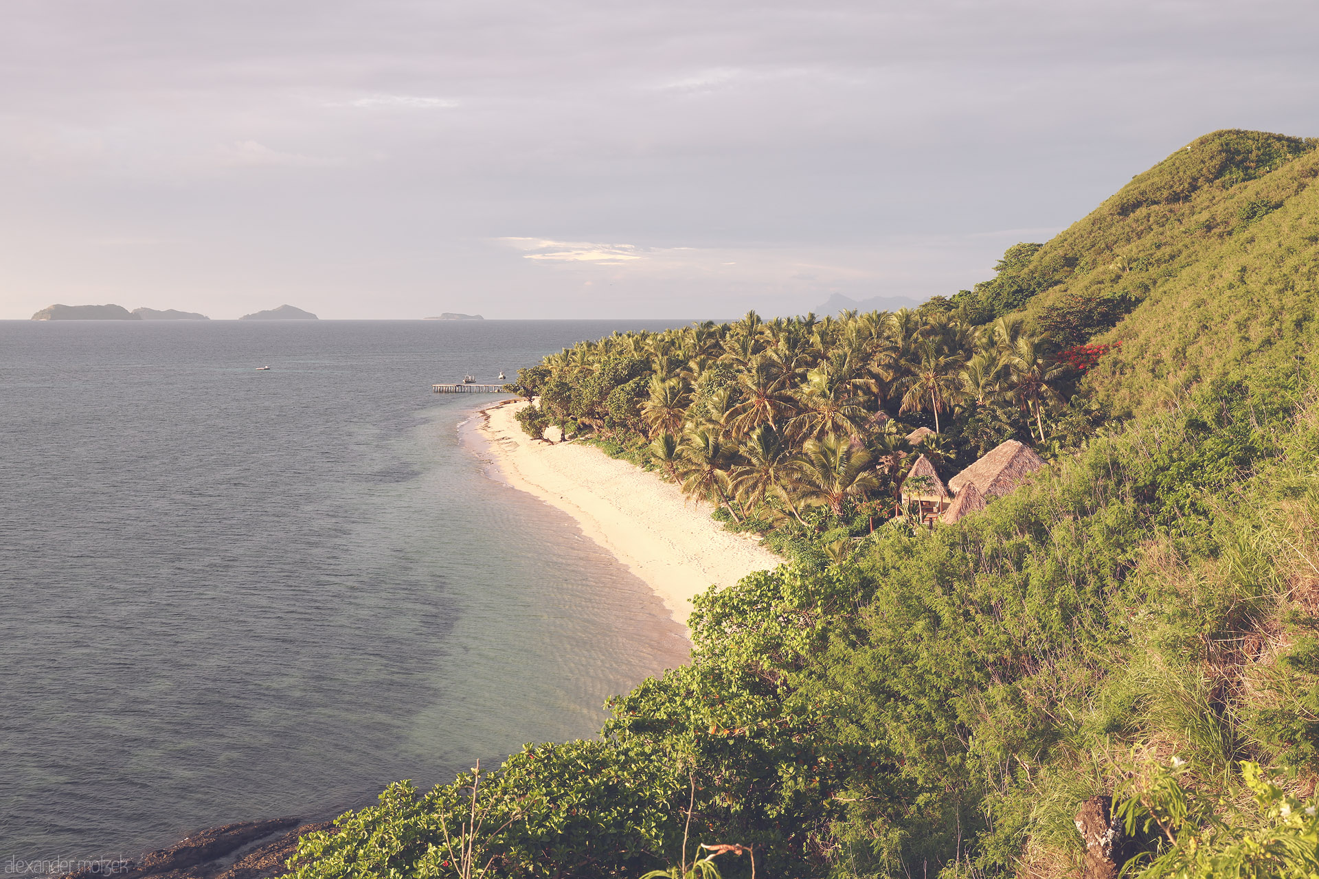 Foto von Palm-lined crescent beach, bures and a small pier beneath lush hills, gazing over calm Pacific waters in Tokoriki, Fiji.