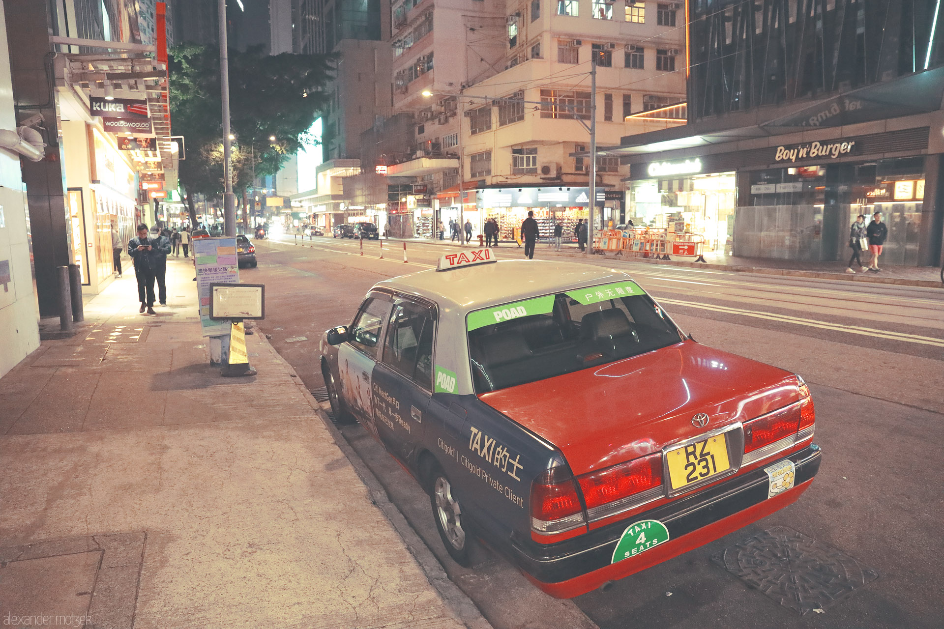 Foto von A classic red Hong Kong taxi parked beside tram tracks, glowing under neon storefronts on a busy Kowloon night street.