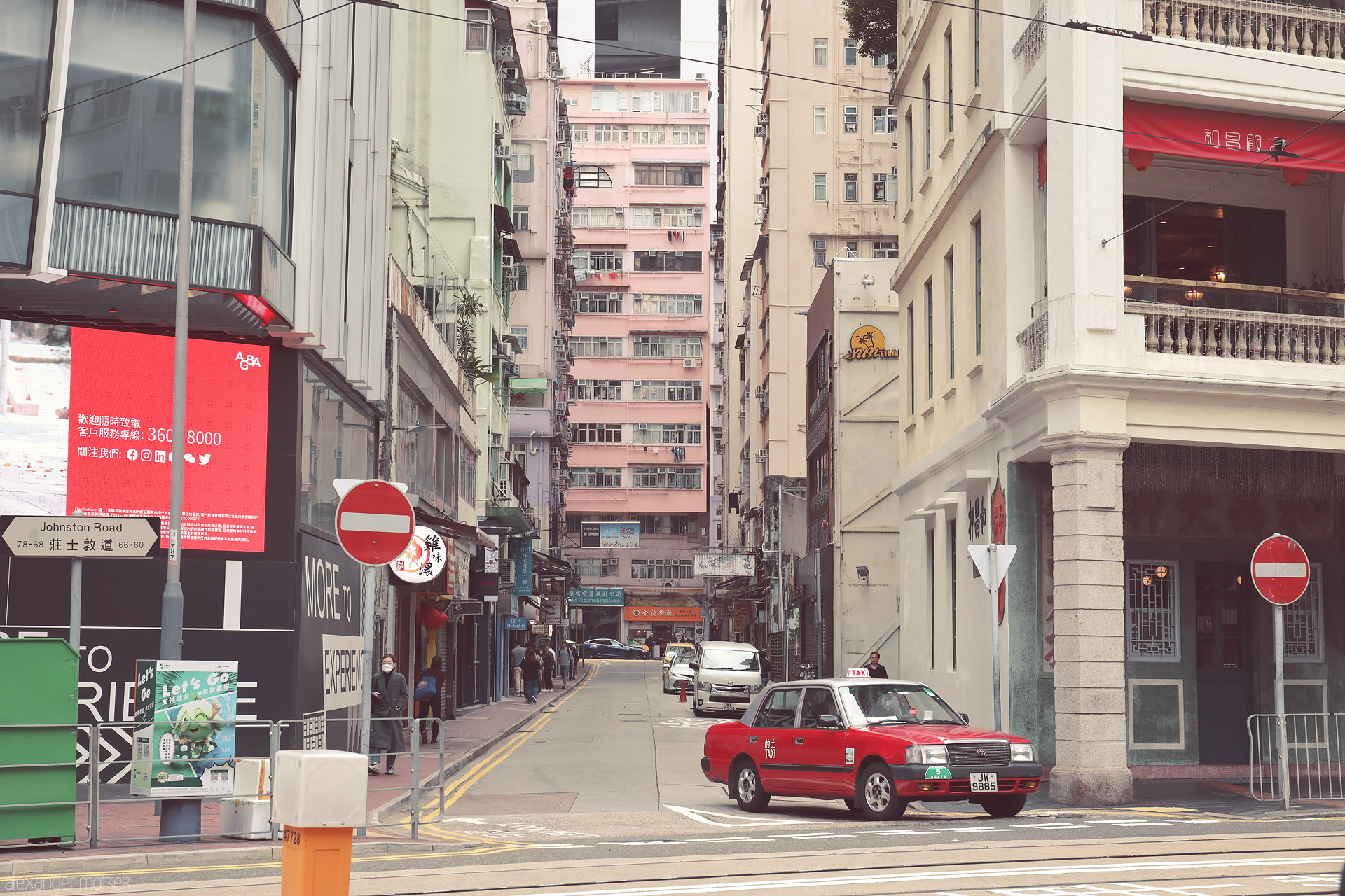 Foto von A classic red Hong Kong taxi turns off Johnston Road into a narrow Wan Chai street, framed by pastel high-rises and shop signs.