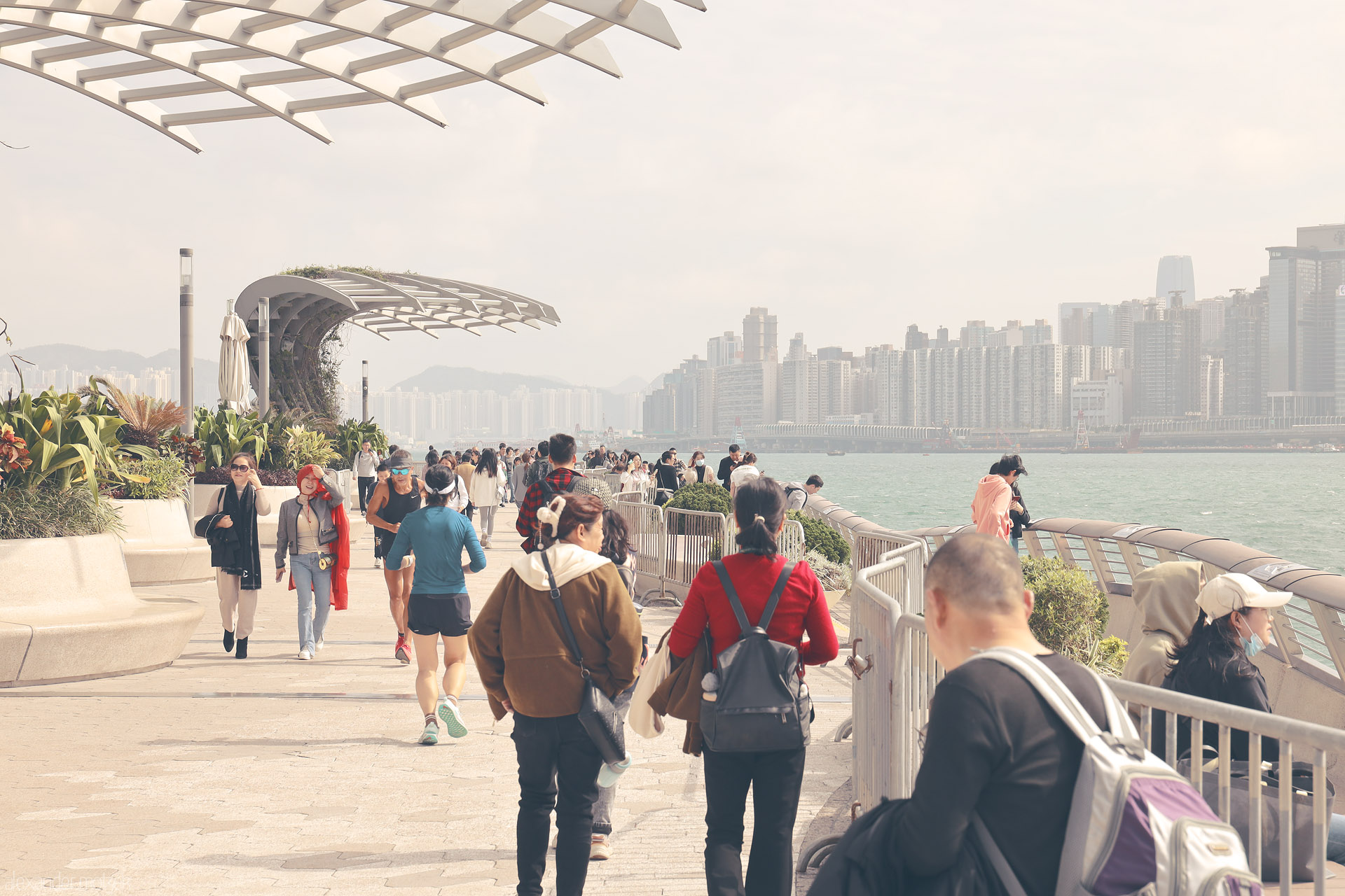 Foto von Crowds stroll the Tsim Sha Tsui waterfront in Kowloon, with Victoria Harbour breeze, curving canopy and a hazy Hong Kong skyline.