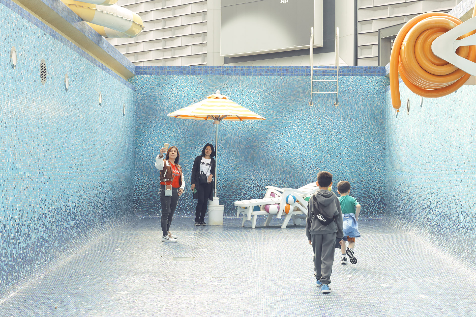 Foto von Empty blue-tile pool in Kowloon, Hong Kong; kids roam past loungers and a striped parasol as visitors pause for selfies.