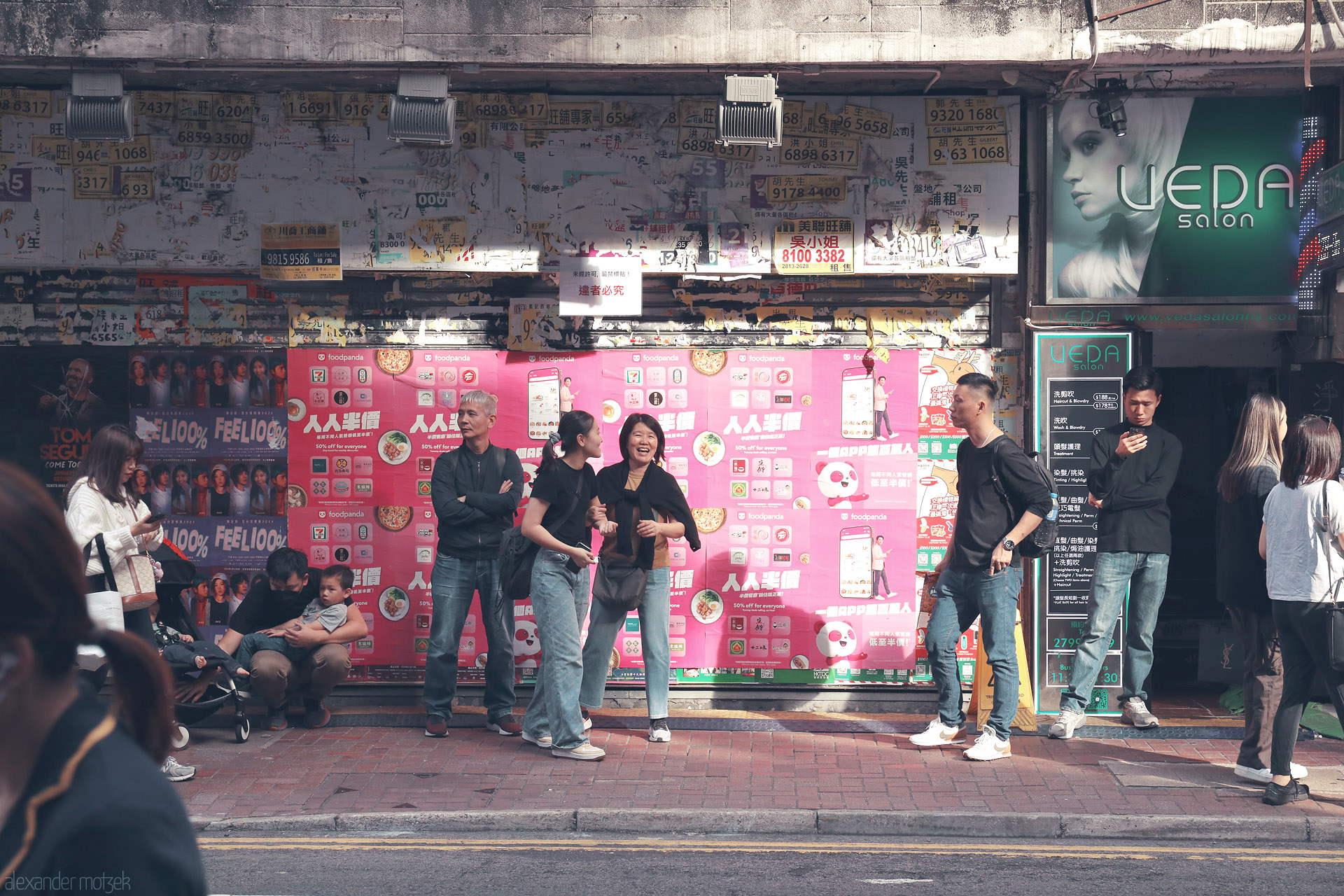 Foto von Wan Chai, Hong Kong: sunlit pavement scene with locals waiting beside pink Foodpanda posters, torn phone-number ads, and a UEDA salon sign.