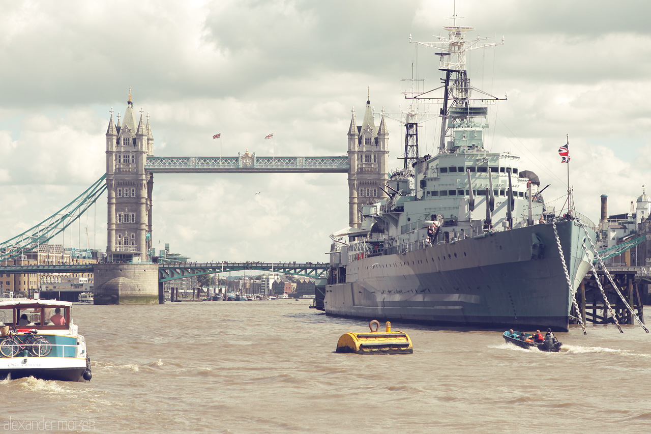 Foto von Iconic Tower Bridge and a majestic warship anchored along the Thames in London. A blend of history and modernity under cloudy skies.