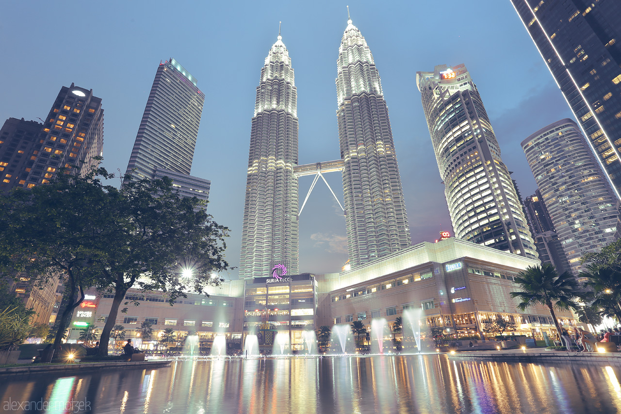 Foto von Dusk envelops Kuala Lumpur as the Petronas Towers sparkle above the cityscape, reflected in tranquil waters below.