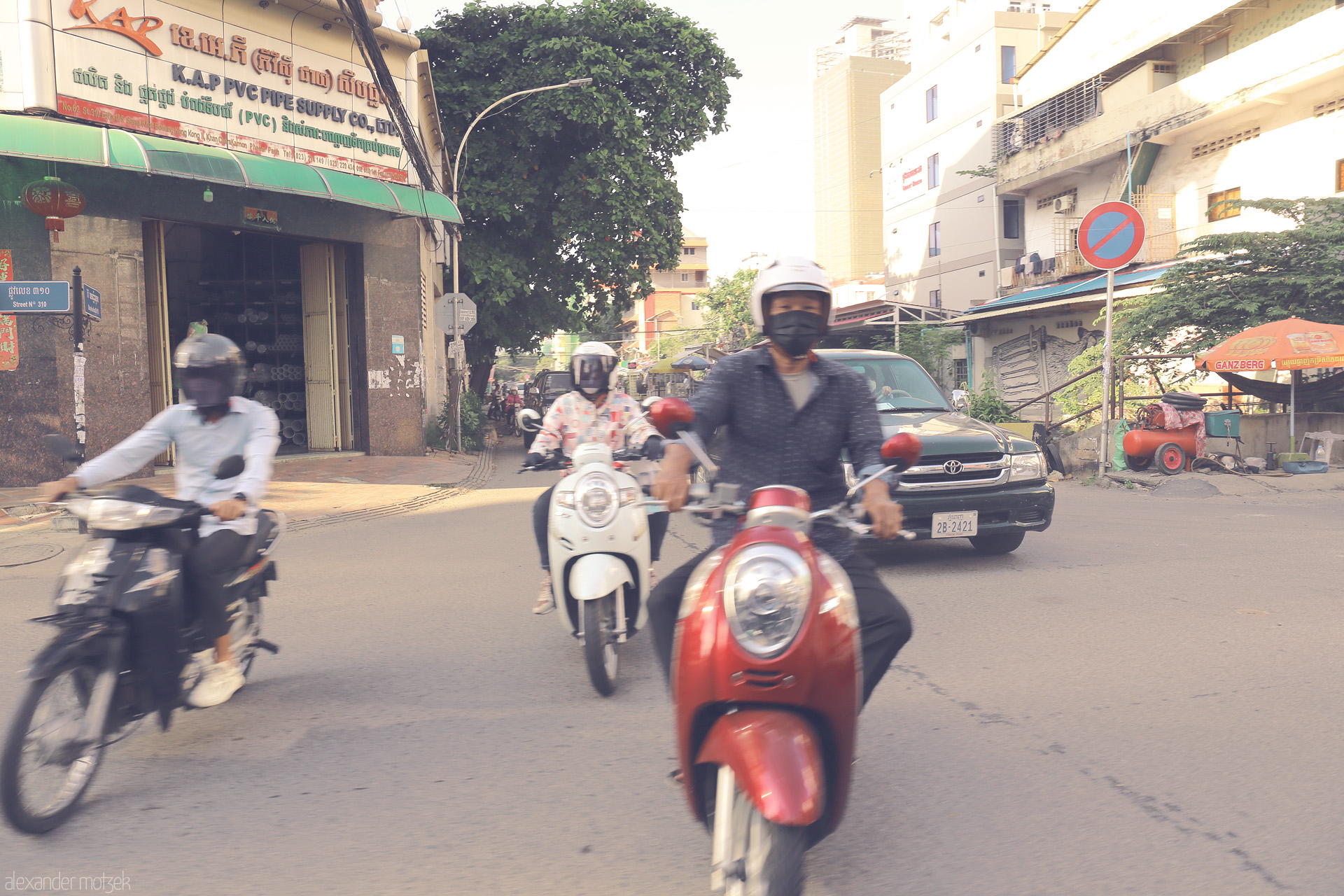 Foto von Morning moto frenzy in Kandal: blurring scooters ride past shops and street signs on sunlit Street 390, Cambodia’s bustling daily rhythm.