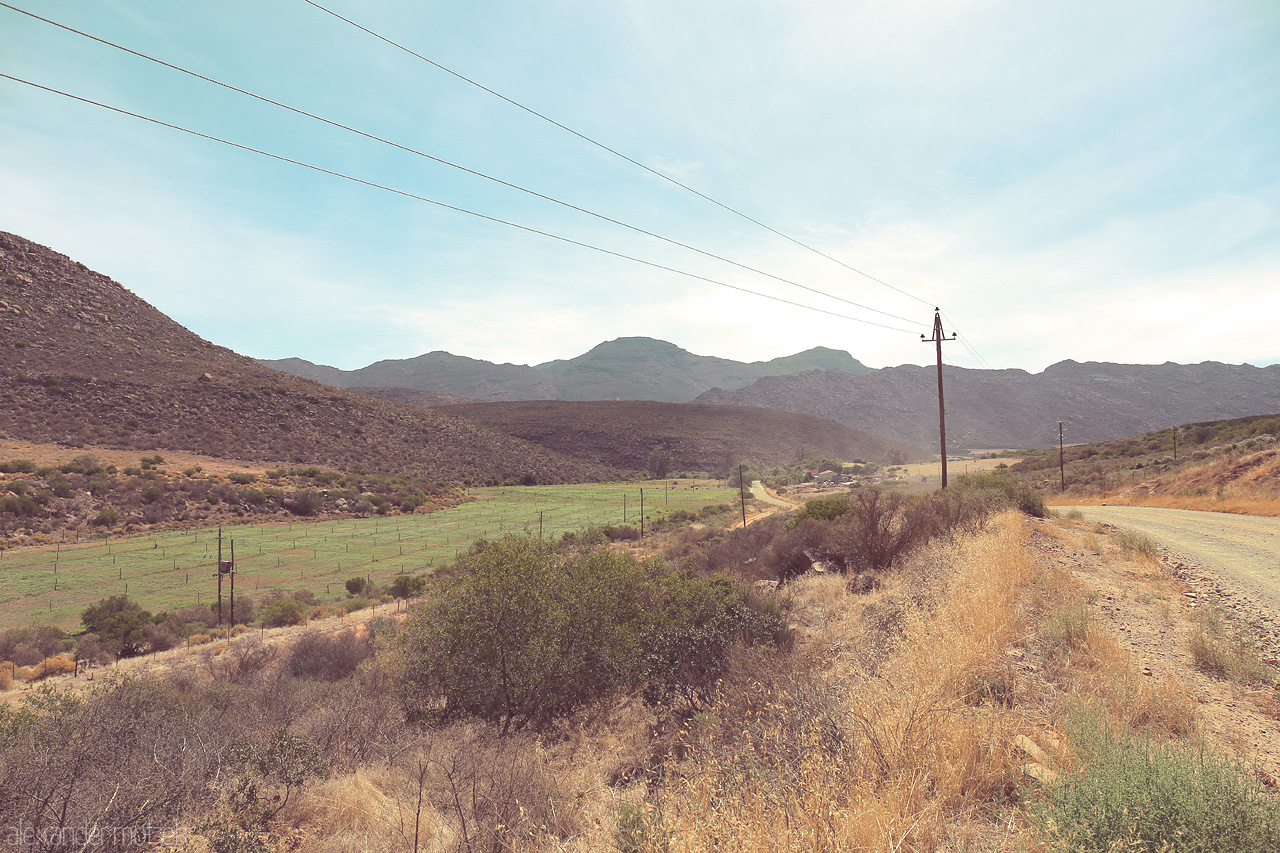 Foto von A winding dirt road through Cederberg's rugged mountains under a vast sky, with fynbos and power lines stretching into the horizon.