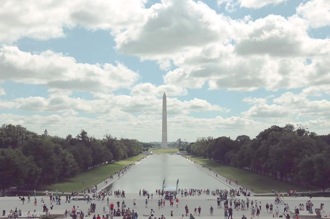 Foto von A serene morning view of the Washington Monument framed by lush greenery and a bustling crowd beneath a canopy of cotton clouds.